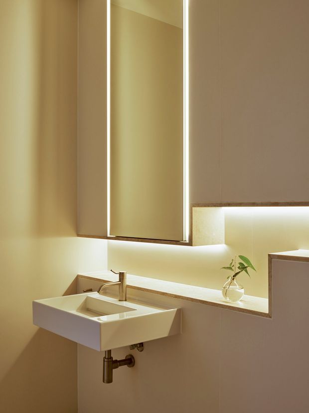 A compact contemporary bathroom vanity with a wall-mounted white rectangular basin, exposed chrome plumbing, and a slim brushed-metal faucet. A large mirrored cabinet with integrated vertical lighting is mounted above the basin, while warm concealed LED lighting illuminates the backsplash and recessed shelf. The space uses pale beige wall finishes, a light stone or engineered-stone benchtop, and minimal decorative greenery.