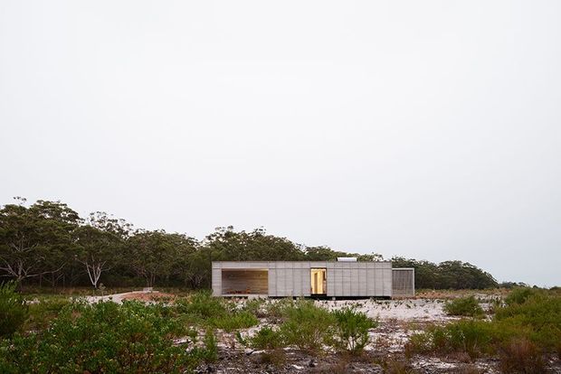 A low, single-storey modern pavilion sits in a sparse coastal or scrub landscape. The building has a long rectangular form, pale grey ribbed or panelled exterior walls, a flat roof, and a warm timber-lined recessed entry or opening. Native vegetation and pale rocky ground surround the structure beneath an overcast sky.