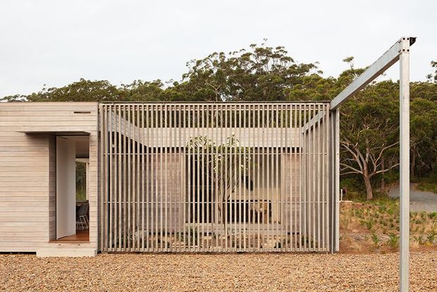 Exterior view of a low, contemporary timber-clad building set in a bushland landscape. A prominent vertical timber batten screen encloses a partially visible outdoor or semi-outdoor space, with a recessed doorway at left and a simple exposed angled steel frame or pergola structure at right. The ground is covered in gravel and native vegetation surrounds the structure.