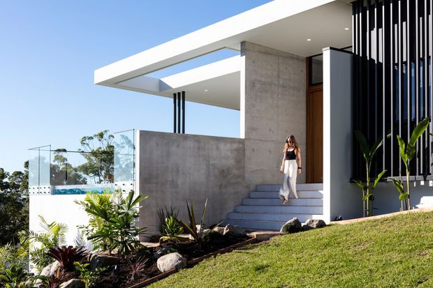Contemporary flat-roofed house exterior viewed from a landscaped approach, featuring exposed concrete walls, crisp white roof planes with a large roof opening, dark vertical facade screens, a timber entry door, glass balustrading, and broad steps rising through tropical planting on a sloped site.