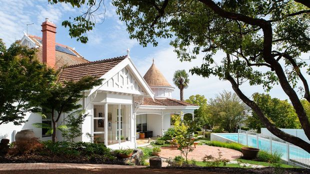 Exterior view of a characterful white-painted residence with ornate gabled detailing, red terracotta tiled roofs, a prominent brick chimney, and a distinctive conical turret roof. The home opens onto a landscaped garden and paved terrace beside a swimming pool, with mature trees framing the scene.