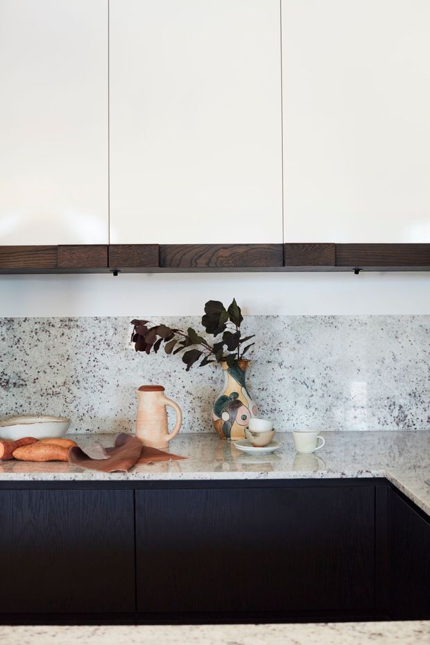 A refined modern kitchen vignette featuring tall off-white upper cabinetry with dark timber trim, dark timber lower cabinetry, and a pale speckled stone benchtop and splashback. Ceramic vessels, cups, bread, and dried foliage add warm organic detail. The compact composition has a restrained, material-focused aesthetic with strong contrast between light surfaces and dark wood joinery.