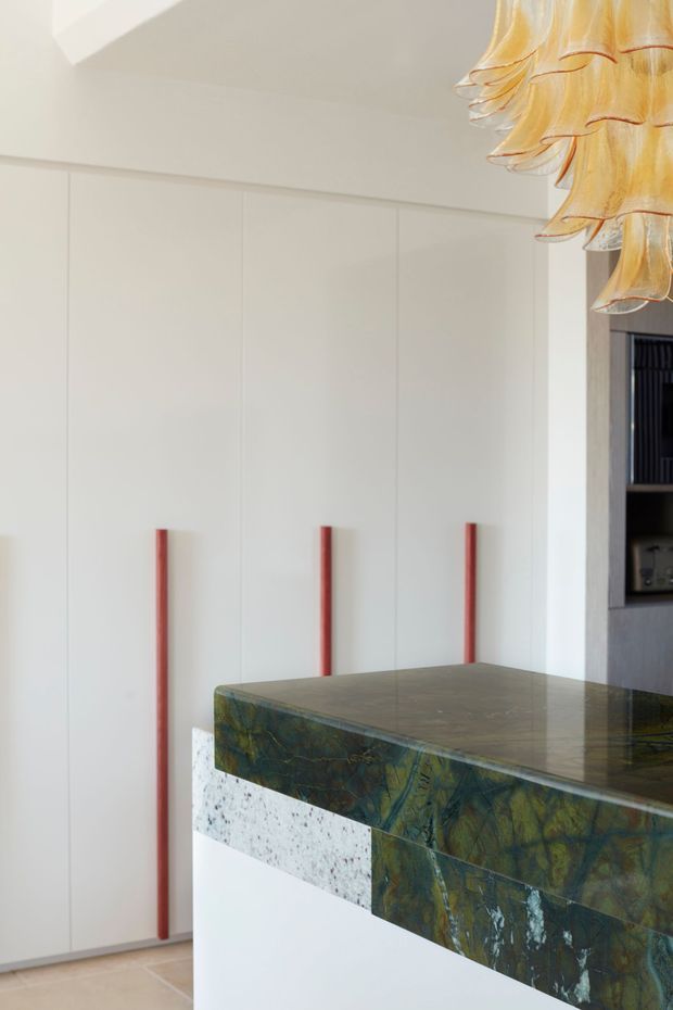 Close-up interior view of a refined contemporary kitchen, dominated by tall white flat-panel cabinetry with slim red vertical handles. A substantial island in the foreground has a dark green, blue and brown veined stone benchtop and matching waterfall-edge sides, contrasted with a white base and a horizontal speckled stone detail. A sculptural warm-toned pendant light hangs at the upper right, with dark built-in shelving and appliances partially visible beyond.