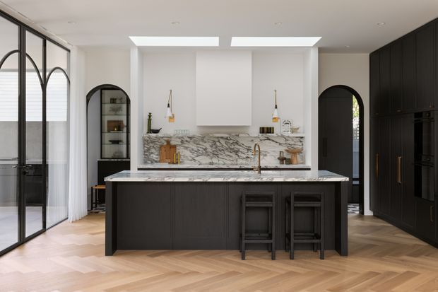 A refined contemporary kitchen with a large dark island, veined marble benchtops and splashback, integrated black cabinetry, arched openings, black-framed glazing, and a warm herringbone timber floor. Two skylights brighten the otherwise monochrome space, with stools positioned at the island.