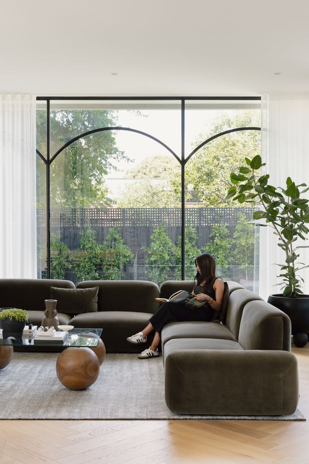 A bright, contemporary living room with a large curved sectional sofa in muted olive-grey upholstery, a low glass coffee table, sculptural wooden round stools, and a large neutral rug. Floor-to-ceiling glazing features distinctive arched black framing, with sheer curtains and a leafy planted garden visible beyond. Pale herringbone timber flooring and a tall indoor potted plant add warmth and texture.