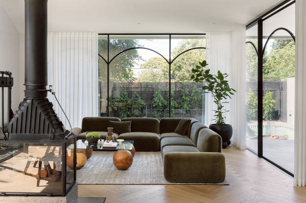 Bright contemporary living room with floor-to-ceiling black-framed glazing, distinctive arched window mullions, white sheer curtains, and views to a planted outdoor area. An olive-green curved sectional sofa surrounds a glass coffee table on a pale rug, while a large black freestanding fireplace with glass panels anchors the left side. Pale herringbone timber flooring, a tall indoor plant, and restrained neutral finishes create a refined, inviting interior.