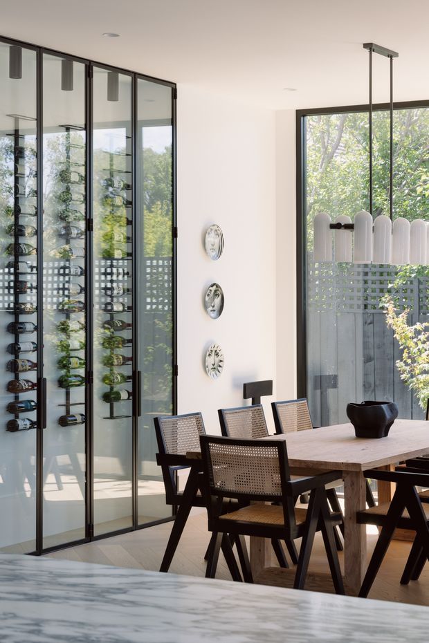 Bright contemporary dining area with a substantial timber dining table and woven cane dining chairs. A full-height black-framed glazed wine display cabinet occupies the left wall, while large black-framed windows and a sculptural multi-arm pendant bring in views of greenery. Decorative circular wall pieces and a marble island or benchtop are visible in the foreground.