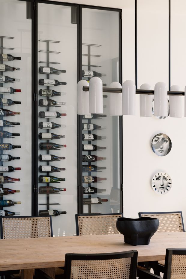 A bright, refined dining area with a long natural timber dining table and woven cane dining chairs. Behind the table, tall black-framed glazed cabinetry displays an extensive collection of wine bottles on slim metal racks. A linear hanging light with repeated white cylindrical shades spans the room, while decorative face plates add visual interest to the white wall.