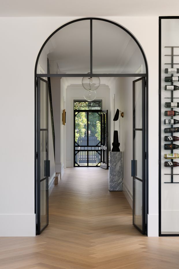 A refined residential entry hall viewed through tall arched black-framed glazed double doors. The hallway has pale herringbone timber flooring, white walls with decorative traditional moulding, black-framed glazing and doors, globe pendant lights, a marble pedestal displaying sculptural objects, and a wine display rack visible along the right wall. A glazed exterior door at the far end opens toward greenery.