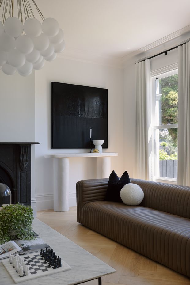Bright, refined living room with white walls, decorative ceiling mouldings, a black fireplace, large abstract artwork, sculptural white console, and a ribbed brown curved sofa. A spherical pendant light hangs overhead, while tall white curtains frame a sunlit window. A marble coffee table with a chess set occupies the foreground.