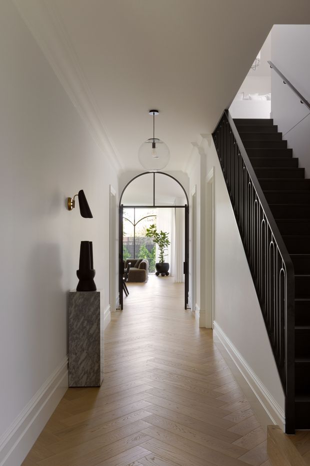 A refined residential hallway with pale herringbone timber flooring, white walls and detailed cornices, leading through a glazed arched doorway into a bright living area. A dramatic black staircase with decorative metal balustrade rises along the right side, while sculptural lighting and a marble pedestal console add contrast.