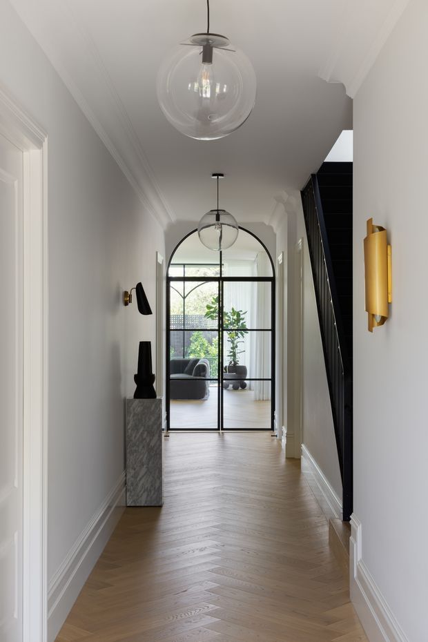 A refined residential hallway with pale grey walls, white decorative cornices and skirting, and a warm timber herringbone floor. A slim marble console with dark sculptural decor sits along the left wall, while black-and-gold wall sconces and two clear globe pendant lights add contrast. At the end, black-framed arched glazing opens visually into a bright living room, with a dark stair or architectural screen along the right side.