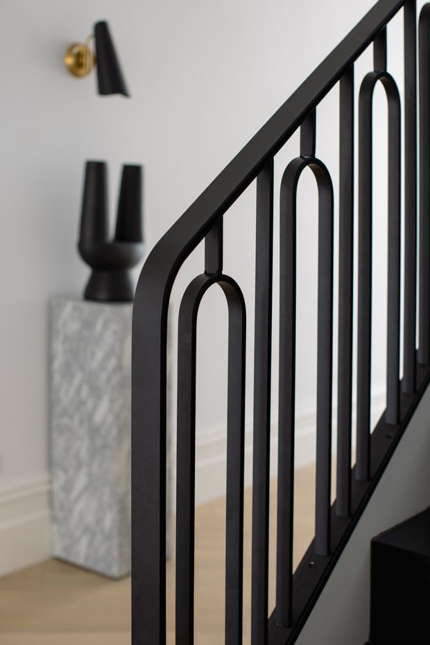 Close-up interior view of a contemporary black metal staircase balustrade with repeated elongated arched vertical details. A pale neutral wall, light timber flooring, marble or stone pedestal, black decorative vessel, and black wall sconce are visible in the background.