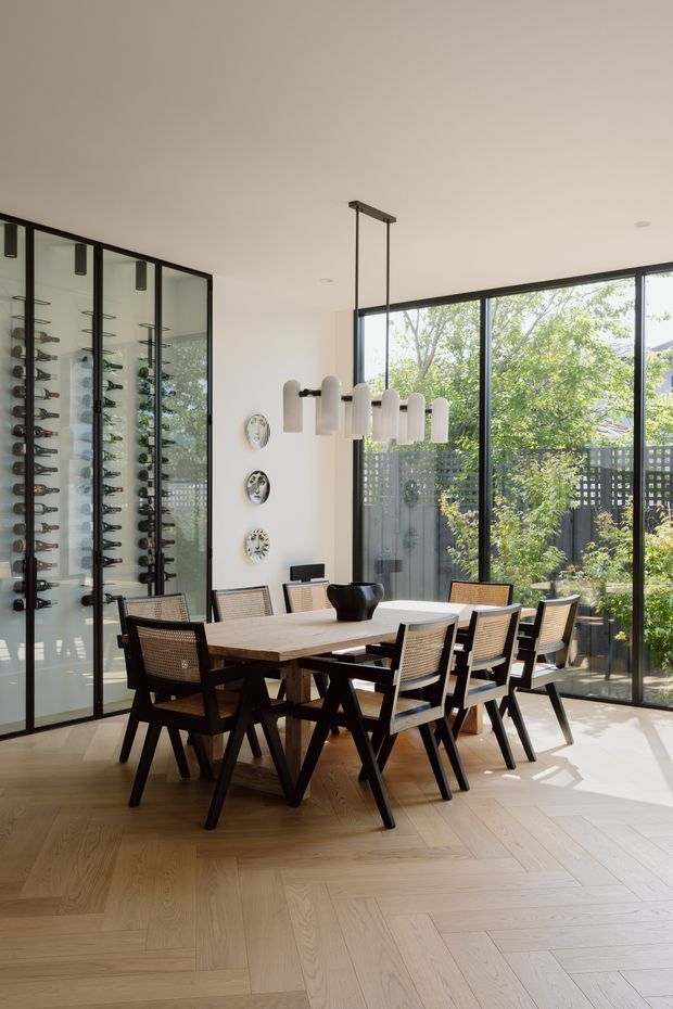 A bright, contemporary dining room with a large timber dining table and black-framed woven dining chairs. A full-height glazed wine display cabinet lines the left wall, while floor-to-ceiling black-framed glazing opens the room to a planted outdoor courtyard. A sculptural white pendant light hangs above the table, with decorative plates arranged on the wall.