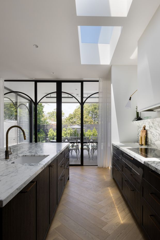 A bright, refined kitchen with two long runs of dark timber cabinetry and white veined stone benchtops. A large island includes an undermount sink and curved brass tapware. Black-framed glazed doors open to an outdoor dining terrace, with sheer curtains at the opening. The herringbone timber floor, arched glazing details, marble-patterned splashback, and two large skylights create a light-filled contemporary interior.