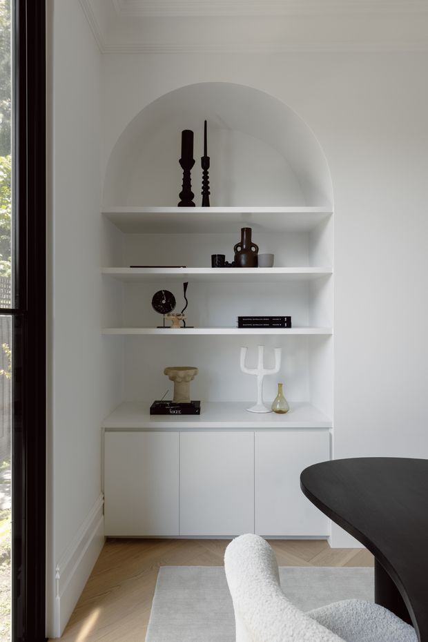 A refined, light-filled dining area featuring a built-in white display cabinet with an arched alcove, open shelves, and closed lower cupboards. The shelves hold curated decorative objects, while a dark rounded dining table, sculptural upholstered chair, pale rug, and timber herringbone flooring complete the space. Tall glazing on the left brings in natural light, and traditional ceiling mouldings add architectural detail.