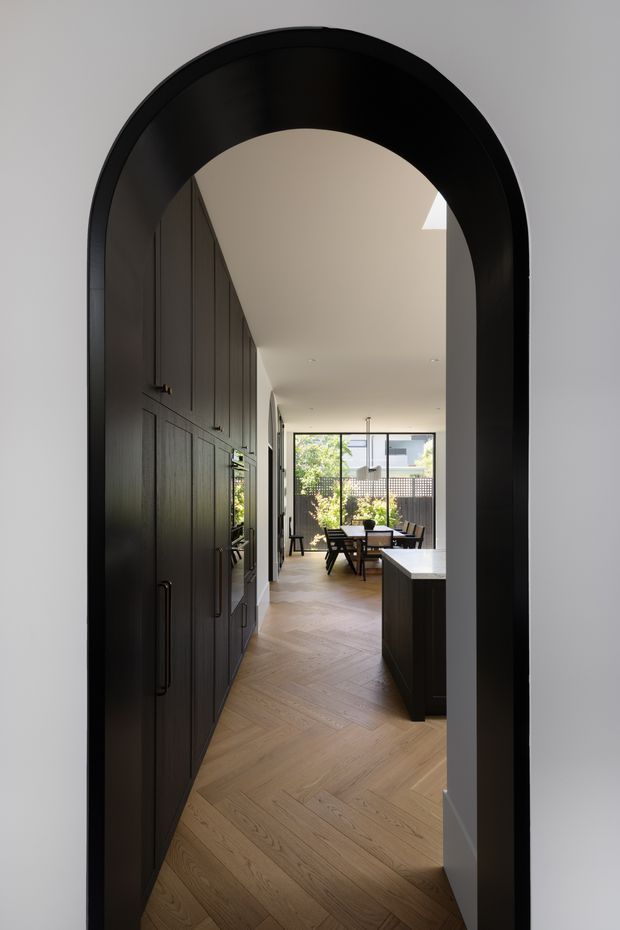 View through a tall arched opening into a refined open-plan interior. Dark built-in cabinetry lines the left side, with a dark kitchen island or joinery element on the right. A light timber herringbone floor leads toward a dining area and large glazed doors or windows at the rear. The palette is restrained, combining black and charcoal joinery, warm timber flooring, white walls and abundant natural light.
