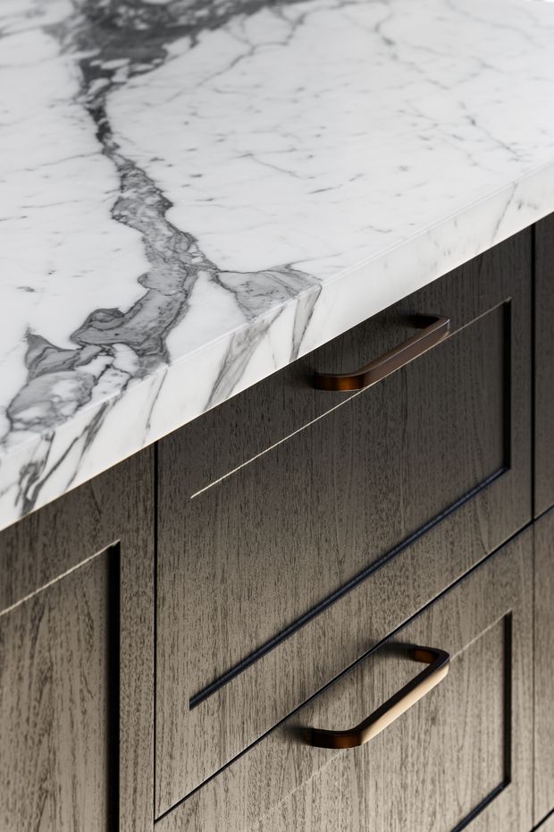 Close-up detail of dark wood-grain built-in cabinetry with slim recessed drawer fronts and bronze-toned bar handles, topped by a thick white marble benchtop with prominent grey veining. The image focuses on refined kitchen or joinery detailing.