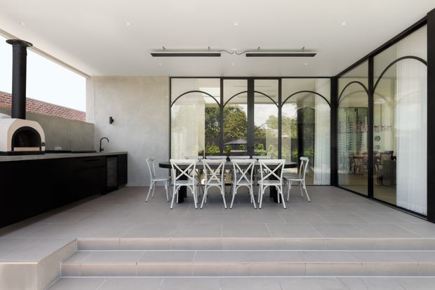 A bright, contemporary indoor-outdoor dining and kitchen area with large arched black-framed glazed partitions. A long white dining table with cross-back chairs sits on a tiled raised platform, while a dark built-in kitchen counter and wood-fired pizza oven occupy the left side. The space opens toward an exterior courtyard through extensive glazing, with a clean neutral palette and strong black framing.