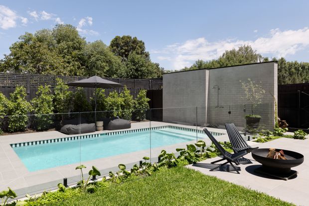 A landscaped backyard pool area with a rectangular turquoise swimming pool, pale tiled paving, frameless glass fencing, dark privacy screens, lush planting, sculptural black outdoor loungers, umbrellas, and a round fire bowl. The scene is bright and contemporary, with a high rendered garden wall and mature trees beyond.
