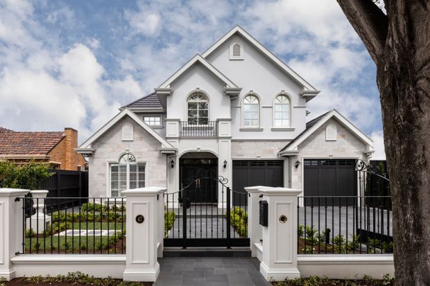 A large, symmetrical multi-storey suburban residence with a white and pale-grey rendered facade, multiple gables, arched windows, decorative trims, balconies, and two dark garage doors. The front property is enclosed by black metal fencing and white masonry pillars, with a paved entry path and landscaped garden beds. A neighbouring brick-tiled roof and mature tree are visible at the edges.