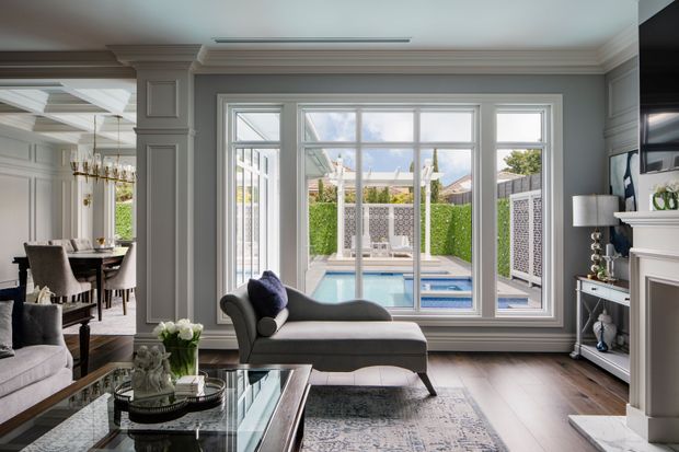 Refined living room with grey-blue paneled walls, decorative ceiling mouldings, large white-framed windows overlooking a swimming pool and landscaped courtyard, and an adjoining formal dining area. The room contains a chaise lounge, sofas, glass coffee table, patterned rug, console cabinetry, fireplace surround and layered classic furnishings.