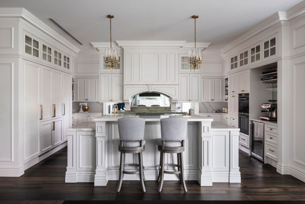A spacious, highly detailed white traditional kitchen with extensive raised-panel cabinetry, integrated appliances, a large central island with two upholstered stools, dark timber flooring, mirrored splashback, and twin brass-and-crystal pendant lights. The room features ornate cornices, pilasters, decorative cabinetry, and a refined, symmetrical composition.