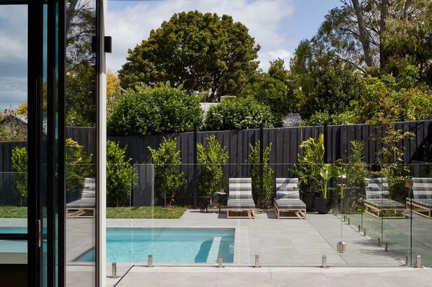 A contemporary outdoor pool courtyard viewed through floor-to-ceiling glazing. The rectangular turquoise pool sits in a pale paved terrace enclosed by frameless glass balustrades and a dark vertical timber privacy fence, with planted screening, outdoor loungers, and mature trees beyond.