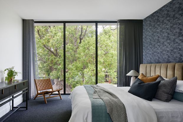 A contemporary bedroom with a large floor-to-ceiling glass opening overlooking leafy trees. The room features a low upholstered bed with layered dark cushions, white bedding and a textured blue-grey runner, a dark textured feature wall, grey curtains, dark carpet, a slim console, and a woven timber lounge chair.