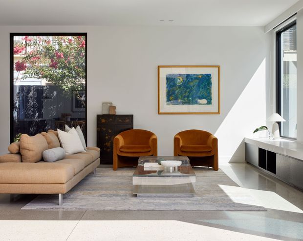 Bright, refined contemporary living room with a large neutral sectional sofa, two sculptural cognac lounge chairs, a transparent glass coffee table, low black-and-white media cabinetry, and a framed blue-green artwork. Full-height glazing and strong sunlight create an airy atmosphere, with a dark timber sideboard and indoor plant adding contrast.