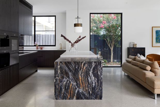 Open-plan contemporary kitchen and living area with dark built-in cabinetry, integrated appliances, a dramatic veined stone island, polished concrete flooring, and a neutral sofa. Large black-framed windows bring in natural light and overlook a planted garden. A sculptural pendant hangs above the island.