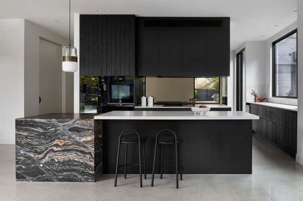 Contemporary kitchen featuring a large dark island with a pale stone benchtop, dramatic veined stone waterfall end panel, black fixed cabinetry, integrated appliances, and two slim bar stools. Full-height glazing and a bright neutral interior create a refined, high-contrast space.