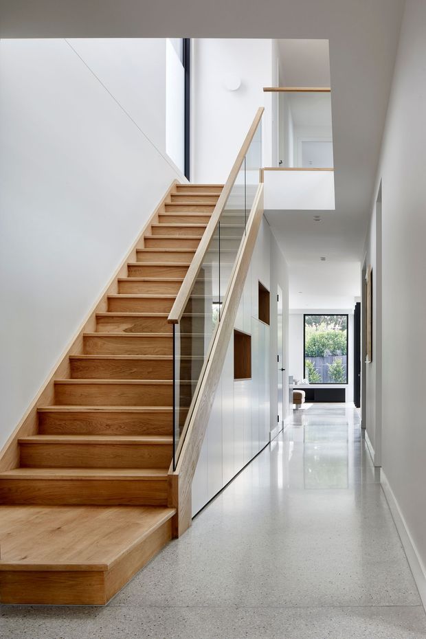A bright contemporary interior hallway featuring a timber staircase with glass balustrade and pale timber handrail. The stair rises beside a tall white wall and opens to an upper landing with simple timber shelving and a clear glass balustrade. The corridor extends toward a glazed rear door or window, with white walls, built-in cabinetry, polished light terrazzo or concrete flooring, and restrained recessed lighting.