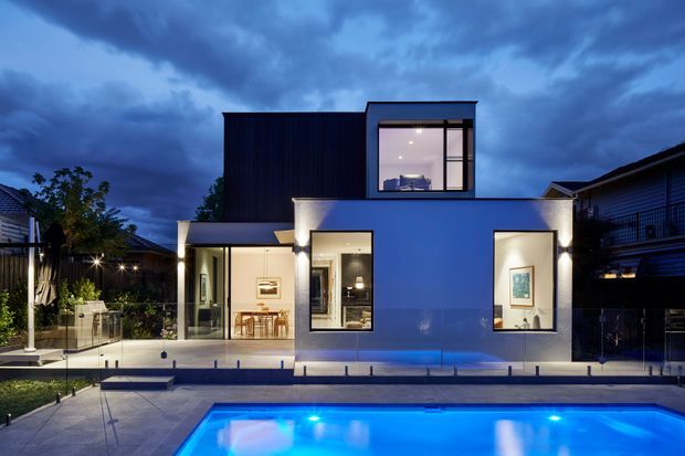 Twilight exterior view of a contemporary two-storey residence with a crisp geometric form, dark upper cladding, pale rendered walls, large windows, and a brightly illuminated swimming pool in the foreground. Landscaped outdoor areas and neighbouring homes are visible around the property.