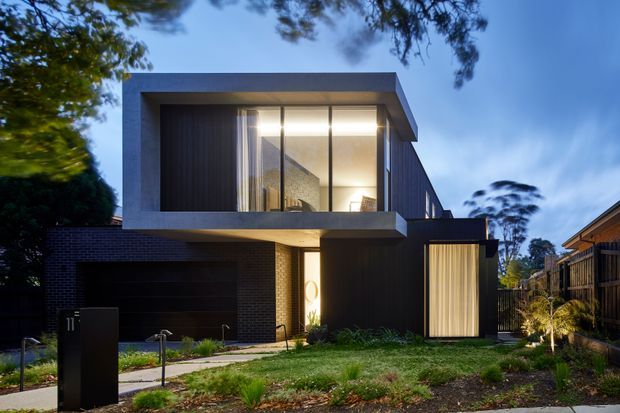 Contemporary two-storey residential facade photographed at dusk, with dark brick and vertical cladding, strong geometric cantilevered upper volume, recessed balcony or living space behind floor-to-ceiling glazing, and an integrated dark garage. Soft landscape planting and warm interior lighting contrast with the dark exterior.