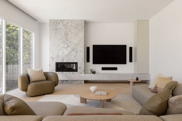 Bright contemporary living room with a large sectional sofa, rounded armchair, timber flooring, marble fireplace surround and built-in television wall. Full-height glazing opens to a leafy outdoor area, while neutral upholstery and warm wood tones create a refined, inviting interior.