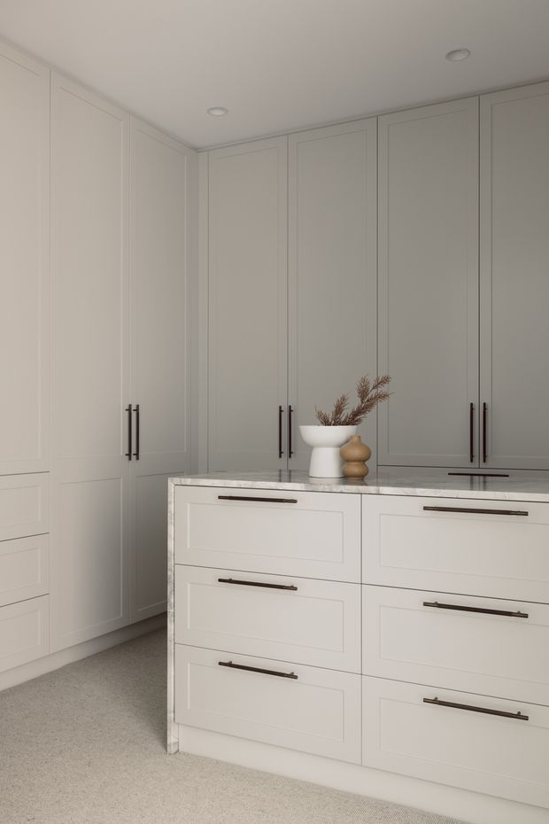 A refined, neutral-toned walk-in wardrobe or dressing room with full-height paneled cabinetry surrounding a central island of drawers. The cabinetry is painted off-white with slim recessed-panel detailing and dark linear handles, topped by a pale veined stone benchtop. Soft recessed ceiling lights, plush light carpet, and minimal ceramic and timber decorative objects create a calm, elegant interior.