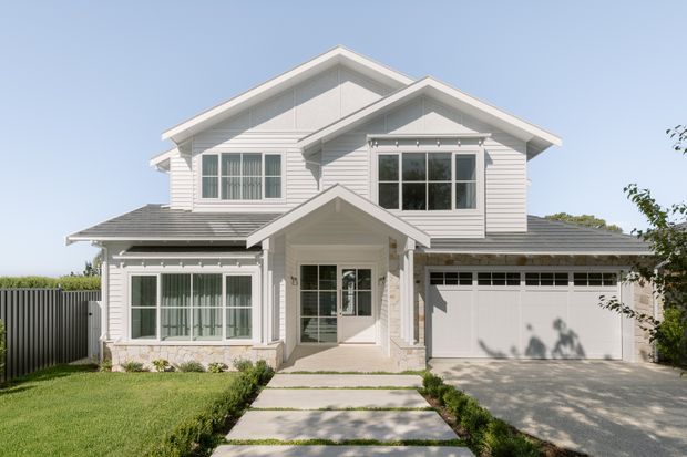 Front exterior view of a two-storey suburban house with a light-coloured weatherboard facade, multiple pitched gables, a covered entry porch, large glazed windows, and an attached double garage. The property has a landscaped front lawn, paved walkway, hedges, and a light grey roof under a clear sky.