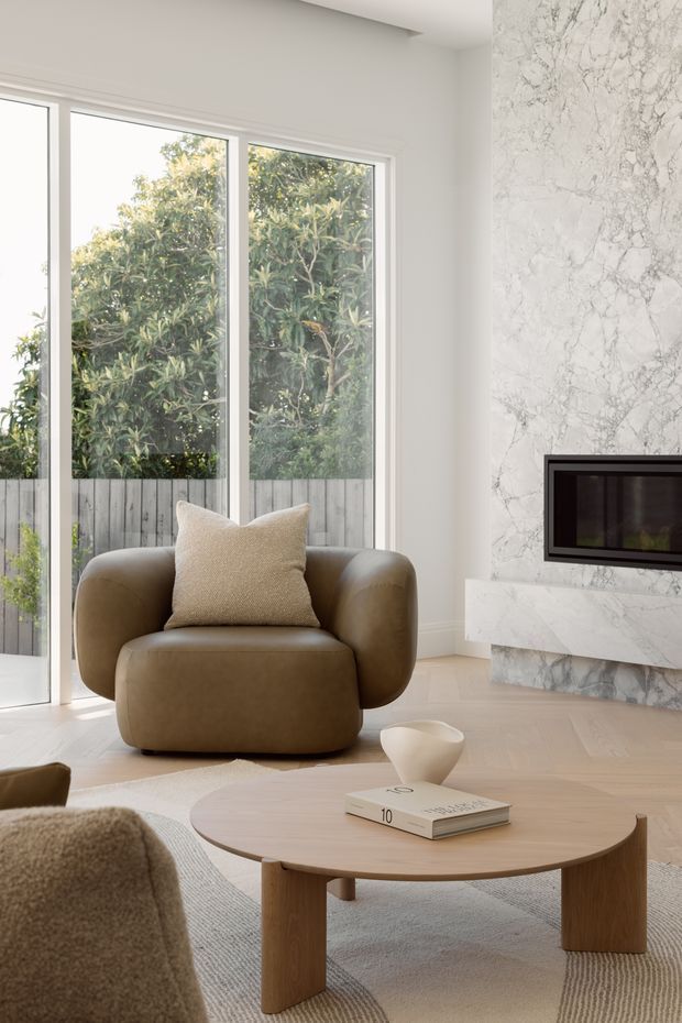 A bright, refined living room with floor-to-ceiling glazing overlooking greenery, a sculptural brown leather lounge chair, a rounded timber coffee table, and a large veined marble fireplace surround with a dark inset firebox. The room uses a warm neutral palette, pale timber flooring, soft textured furnishings, and minimal contemporary styling.