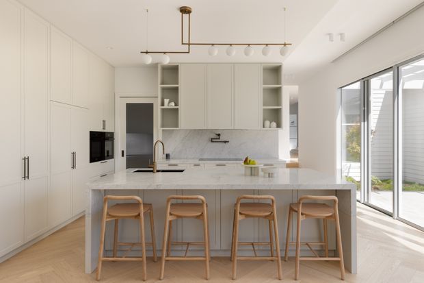 Bright contemporary kitchen with extensive off-white built-in cabinetry, a large white marble-look island with four timber bar stools, integrated appliances, open shelving, and a linear pendant light. Light timber flooring and full-height glazed doors connect the kitchen to an outdoor area, creating an airy, refined space.