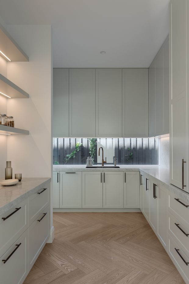 A bright, compact galley-style kitchen with pale grey-green shaker cabinetry arranged along three sides. The room features extensive built-in cabinetry, open illuminated display shelving on the left, a light stone benchtop, a stainless-steel sink with a bronze-toned gooseneck tap, and a narrow horizontal window above the sink looking onto a private planted courtyard. The floor is finished in light timber laid in a herringbone pattern. Dark elongated handles provide contrast against the otherwise soft neutral palette.