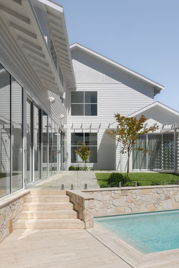 A bright contemporary residential courtyard viewed from a paved pool terrace. The white multi-winged house has horizontal weatherboard cladding, broad glazed openings, a pitched roof with deep eaves, and a covered glass-sided outdoor area. Pale stone steps and paving lead past a low irregular stone retaining wall to a turquoise swimming pool, with a small landscaped courtyard and young trees.