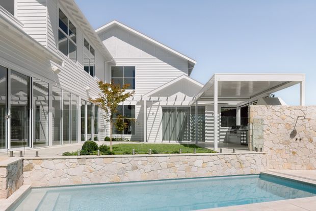 A bright contemporary coastal-style residence viewed from the pool courtyard. The building has white horizontal weatherboard cladding, pitched gabled roofs, extensive glazing, and a covered outdoor entertaining area with a stone-clad wall and outdoor kitchen. A rectangular swimming pool and landscaped courtyard occupy the foreground.