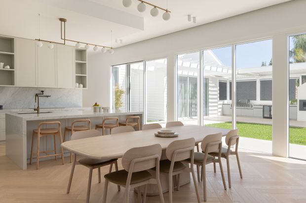 Bright open-plan kitchen and dining area with a large light-wood dining table and upholstered chairs in the foreground. The kitchen has pale cabinetry, open shelving, a marble-look backsplash, a stone island with timber stools, and contemporary spherical pendant lighting. Floor-to-ceiling glazing opens onto a sunny courtyard and connects the space to an outdoor covered area.