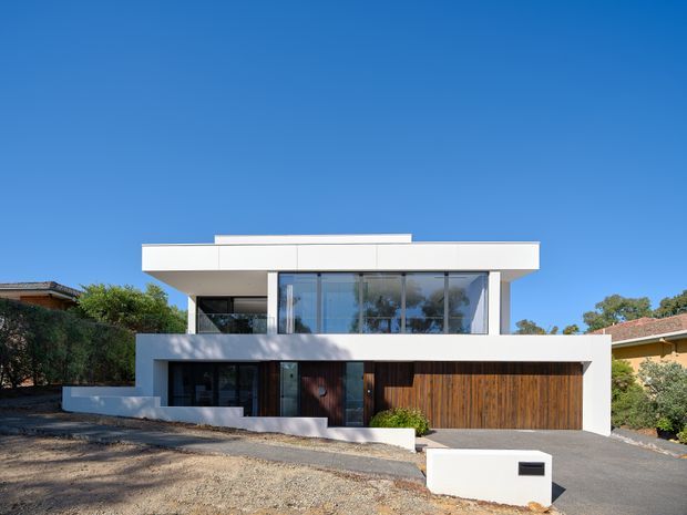Front exterior view of a two-storey contemporary residence with a strong rectilinear composition, broad flat roof overhang, white rendered facade, extensive upper-level glazing, dark window frames, and a warm timber-clad entry and garage door. The home sits on a suburban site with a driveway and low garden planting under a clear blue sky.