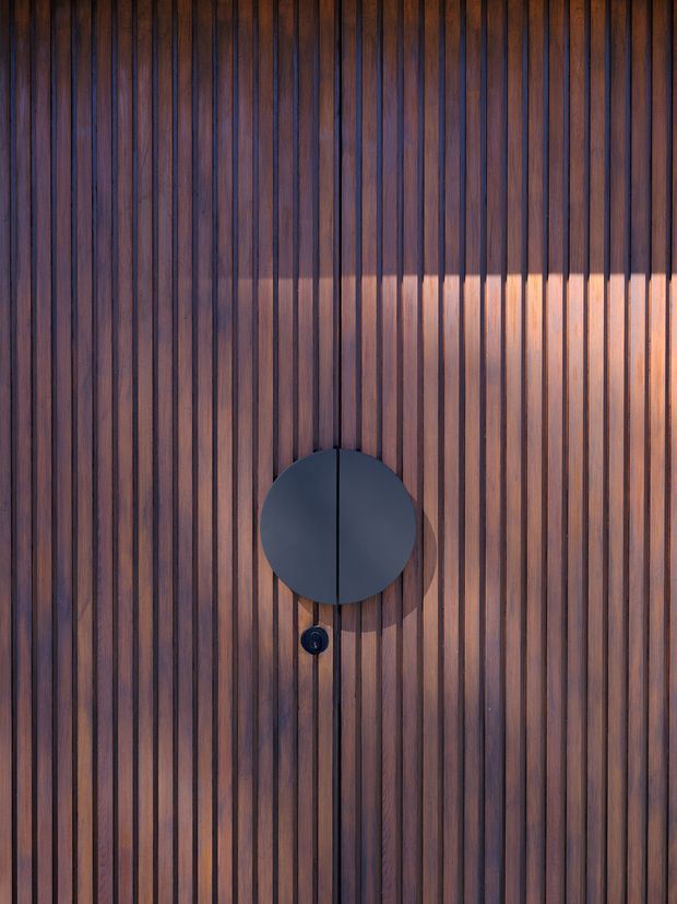 Close-up architectural detail of a vertically slatted dark timber door or wall panel. The narrow, evenly spaced timber battens create a strong linear rhythm, with a large matte black circular door pull or feature plate and a small black lock below. Warm sunlight highlights the natural wood grain and casts a horizontal band of light across the surface.