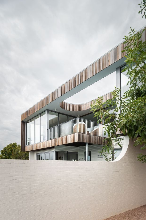 Exterior view of a contemporary multi-level residence with extensive glazing, vertical timber cladding, a projecting flat roof, curved roofline cut-out, and a balcony with vertical metal balustrading. A pale rendered brick boundary wall and landscaped planting sit in the foreground.