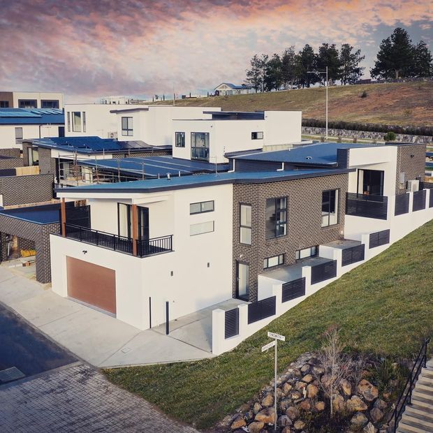 Aerial view of a contemporary multi-storey residential development on a sloped suburban site. The homes feature flat blue-toned roofs, crisp white rendered walls, dark grey brickwork, black metal balcony railings, large windows, and integrated garages. Terraced landscaping, retaining walls, and neighbouring modern homes are visible around the development.