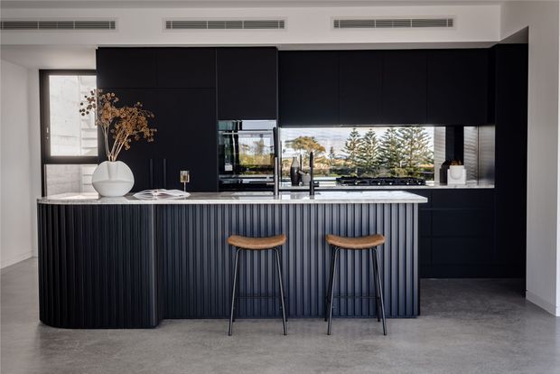 A sophisticated contemporary kitchen with a large black island featuring vertically fluted cabinetry, a pale stone benchtop, matching dark full-height cabinetry, integrated appliances, and two timber bar stools. A wide horizontal window above the rear counter brings in natural light and overlooks trees. The room has a polished concrete floor, white walls, and linear ceiling air vents.