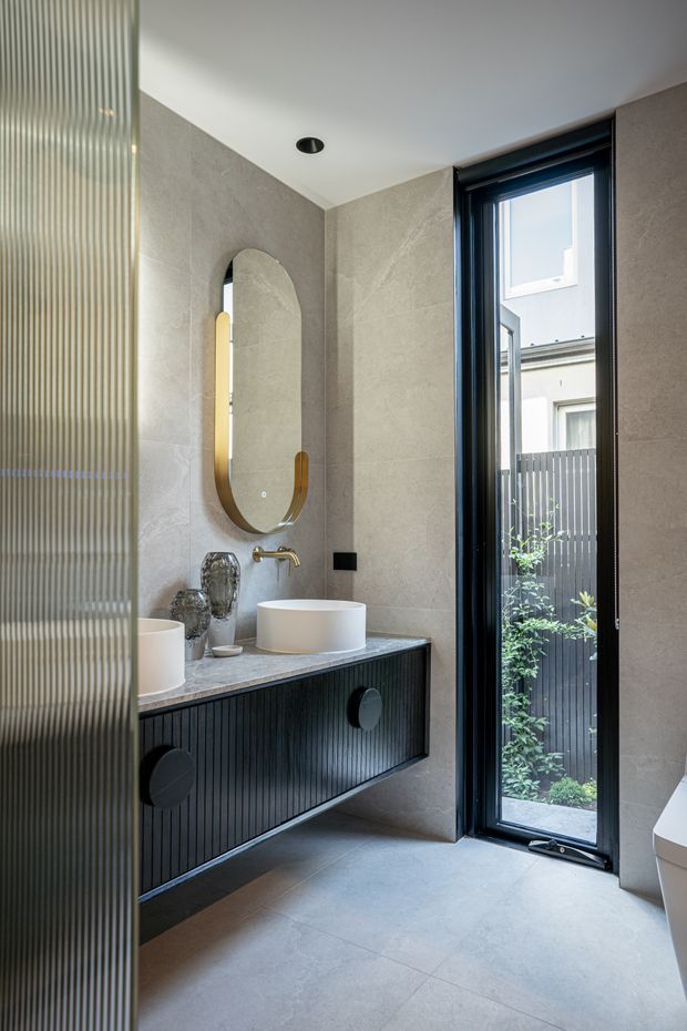 A contemporary bathroom with large grey stone-look tiled walls and floor, a floating dark fluted vanity with a grey stone benchtop, two round white vessel basins, and a brushed brass wall-mounted tap. An elongated arched mirror hangs above the vanity, while a tall black-framed glazed door opens to a small private courtyard. A ribbed translucent glass partition partially screens the room.